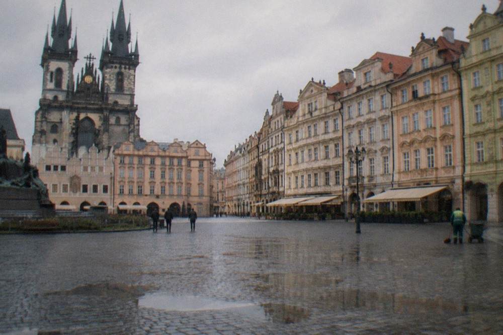 Una plaza empedrada en la Ciudad Vieja de Praga con fachadas barrocas de colores pastel y una torre de piedra al fondo