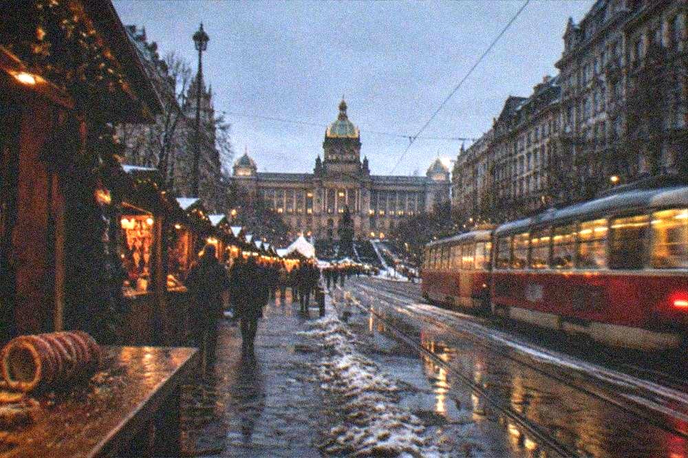 Plaza abierta en Praga al anochecer con puestos de madera de mercado y un árbol iluminado frente a edificios históricos