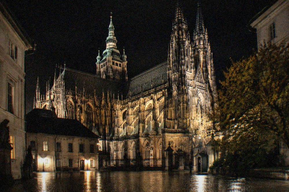 Floodlit cathedral spires and castle walls seen from a dark courtyard
