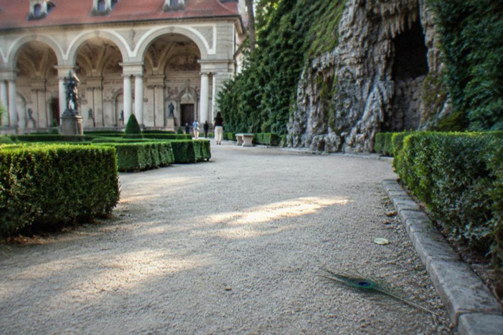 Formal baroque garden with clipped hedges, gravel paths and a stone fountain wall under a blue summer sky