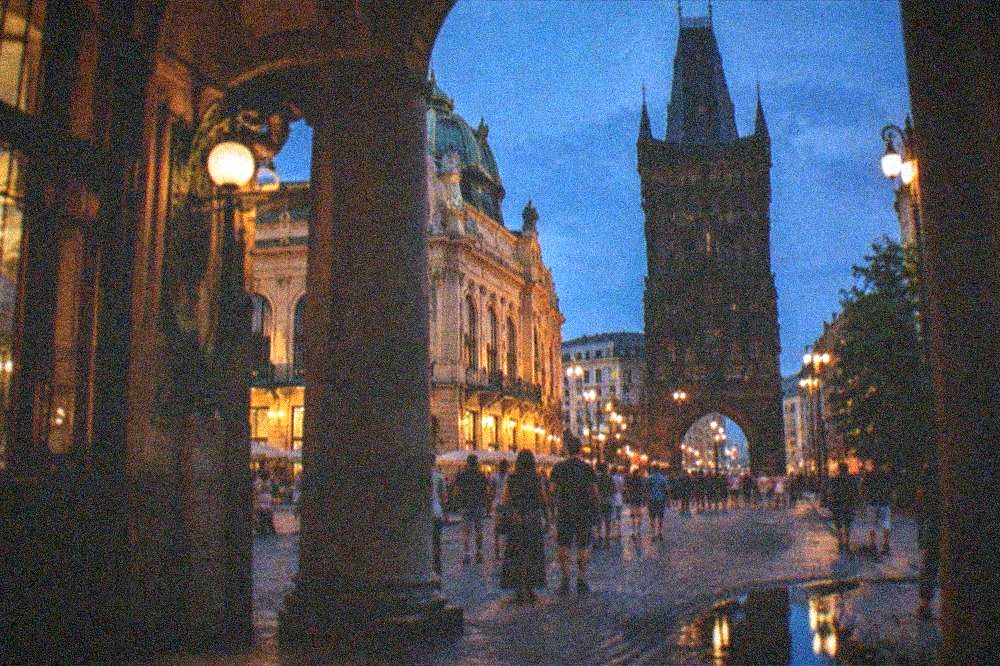 Illuminated historic square at dusk with crowds strolling between lit facades and a dark blue summer sky