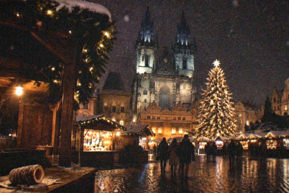 A Christmas market square at night with wooden stalls, a lit tree and gothic towers behind
